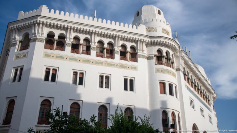 Large white corner building with upper arcade, crenellated roofline, rounded turret, and tall windows.