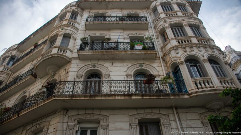 Low-angle view of an ornate corner apartment building with iron balconies, rounded bays, arched windows, and carved stone details.