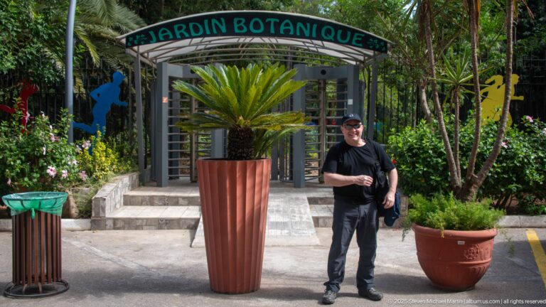 Entrance marked Jardin Botanique with a metal turnstile, potted cycad, colorful figures, and a visitor standing nearby.