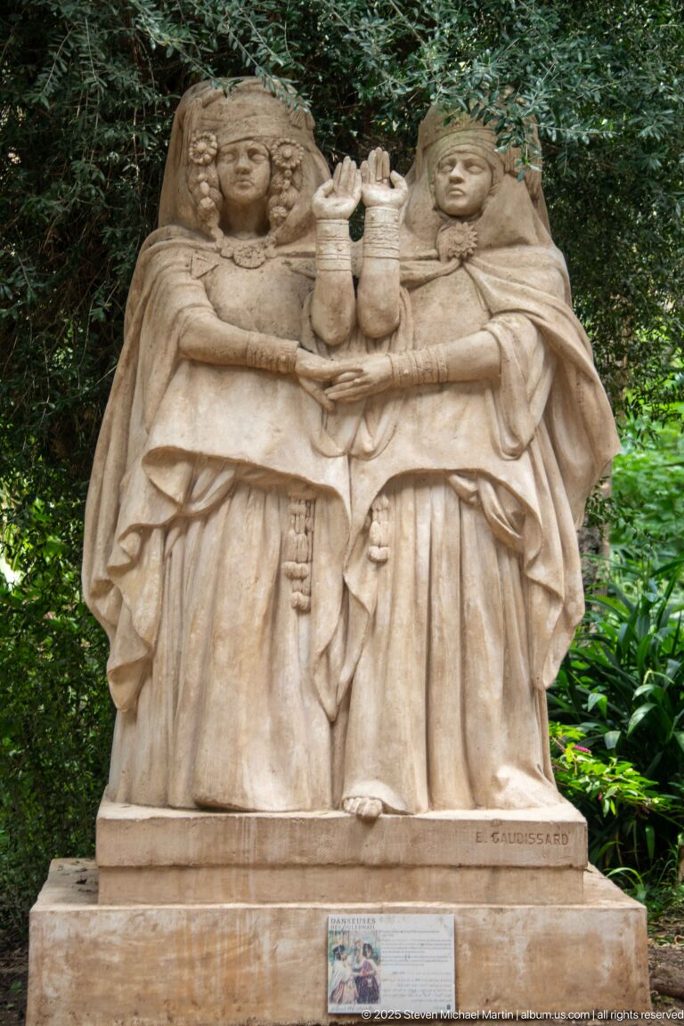 Pale stone sculpture of two robed figures standing together and holding a central vessel or object against dense greenery.