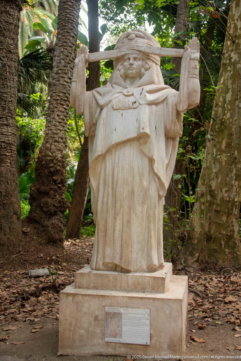 Standing pale stone figure in a robe and head covering, holding a long horizontal staff or beam in a wooded garden.