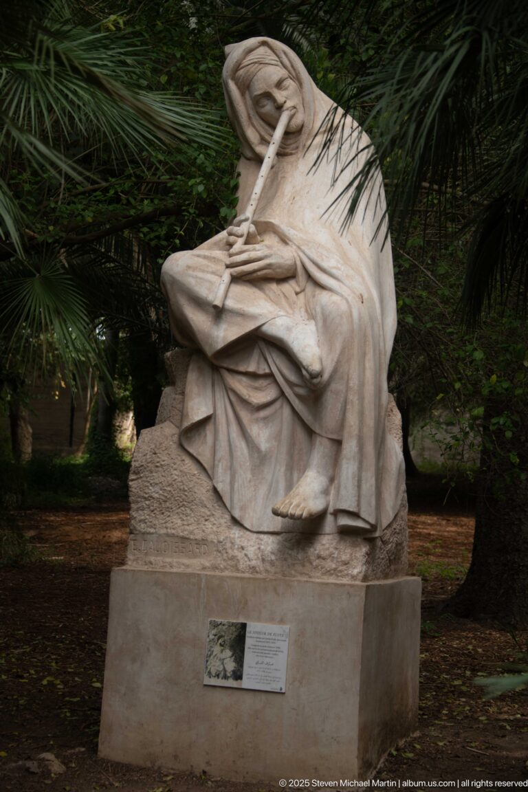 Seated pale stone figure in draped robes playing a long transverse flute beneath palm foliage.
