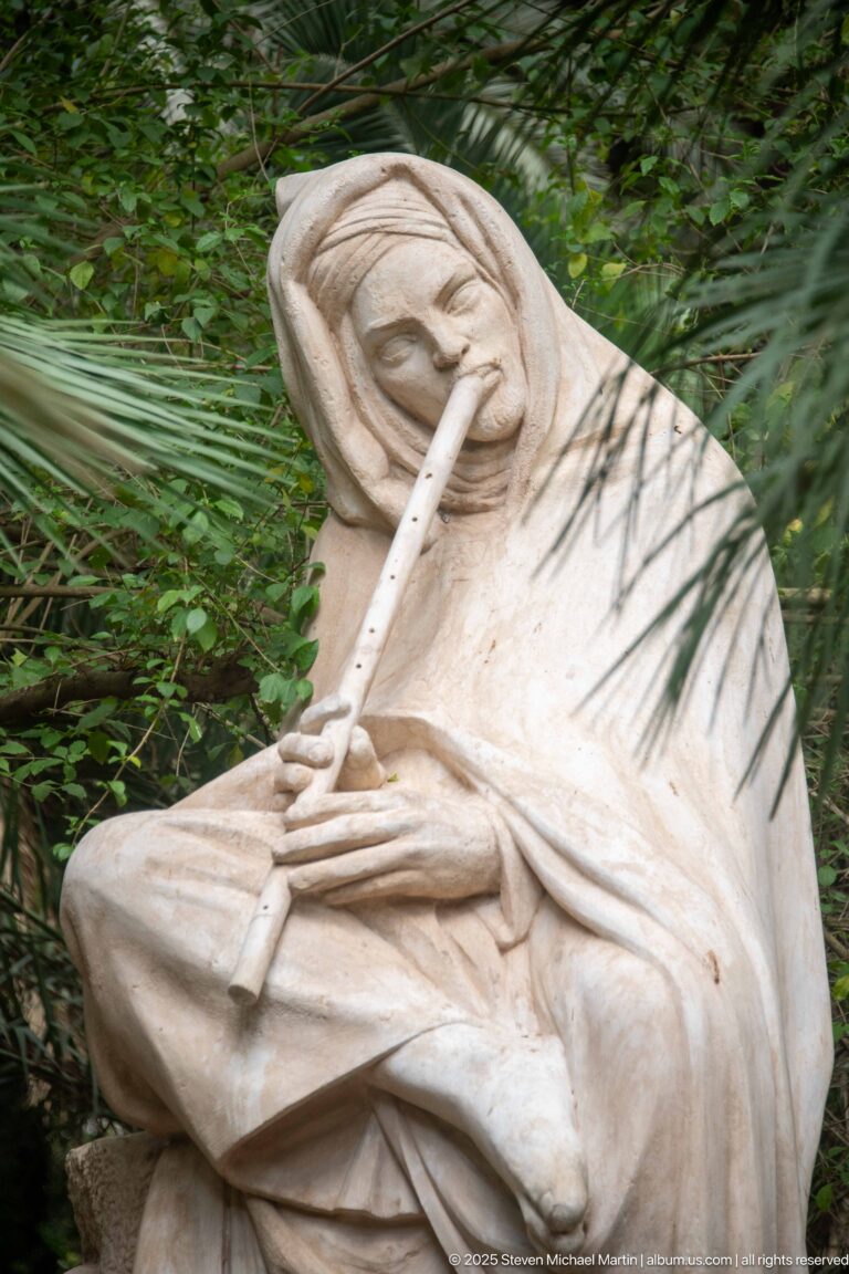 Seated pale sculpture playing a long flute, partly screened by palm-like foliage in the garden.