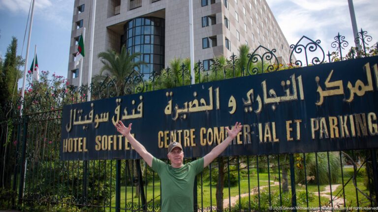 Visitor beneath a bilingual Arabic-French sign reading Hôtel Sofitel and Centre Commercial et Parking beside a hotel building.
