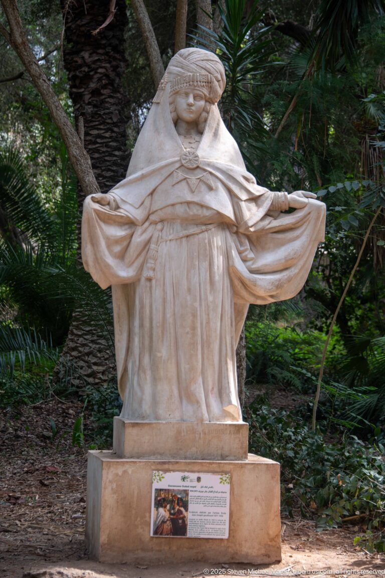 Standing pale female sculpture spreading wide folds of drapery, set on a pedestal among palms and greenery.