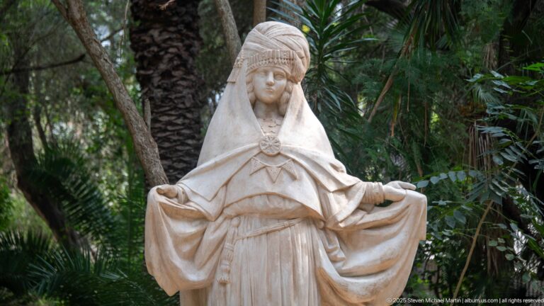 Close view of a pale robed female sculpture with tall head covering and gathered fabric held in both hands.