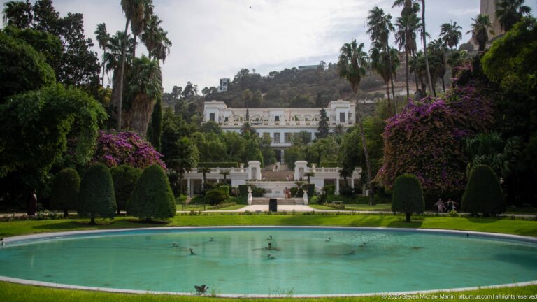 Oval pool and formal garden axis leading toward a white multi-story building on the hillside.