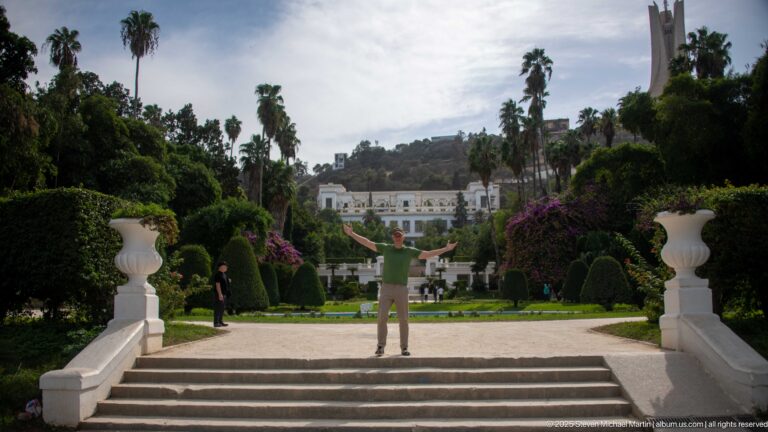 Visitor with raised arms on broad white steps overlooking the formal garden axis, palms, topiary, and distant building.