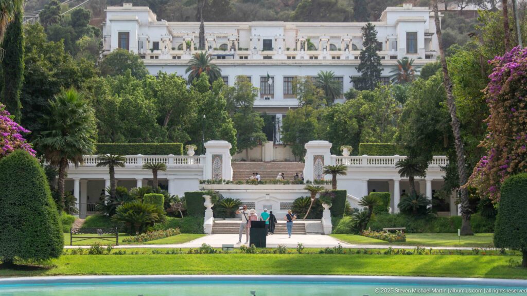 Telephoto view of terraced formal gardens, white balustrades, palms, visitors, and a large white building beyond.