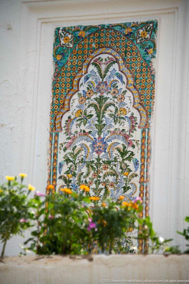 Colorful floral ceramic panel in a white wall, bordered by blue-orange patterns and partly screened by flowers.