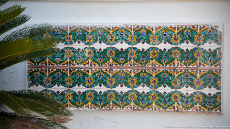 Rectangular wall panel of repeating blue, turquoise, yellow, and orange floral tiles, partly crossed by palm fronds.