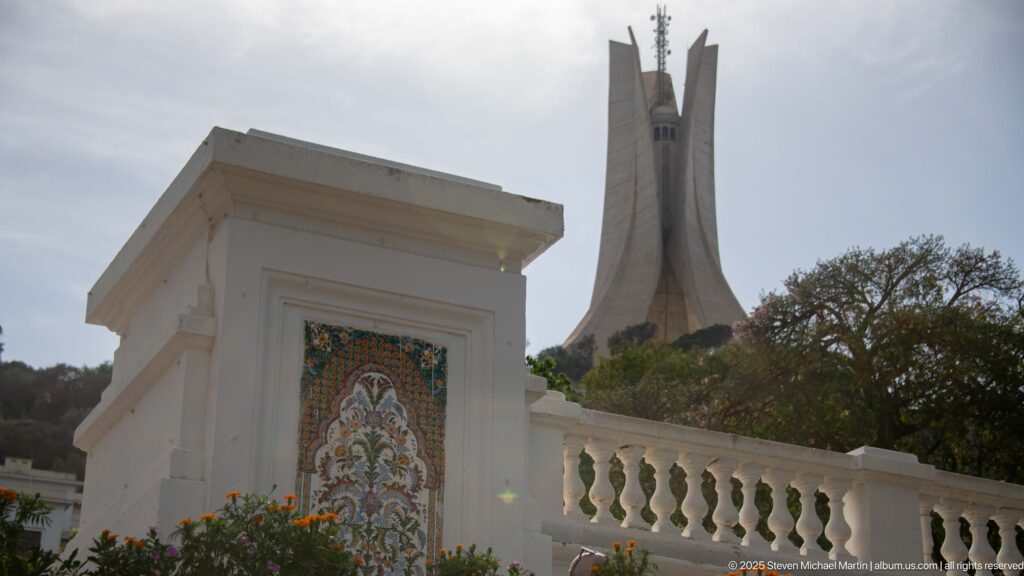 White garden wall with floral tile panel in front of the three-fin Martyrs’ Memorial on the hillside.