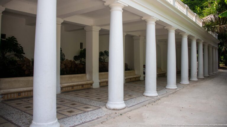 Long white colonnade with round columns, patterned floor tiles, balustraded roofline, and shaded plant displays.
