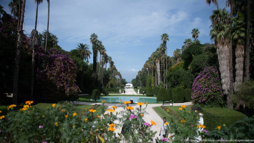 Long formal water channel between lawns and rows of palms, with flowers in front and a white fountain beyond.