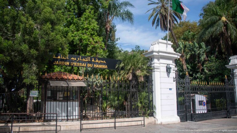 Gated Jardin d’Essai du Hamma entrance with Arabic-French sign, white piers, Algerian flag, and palms.