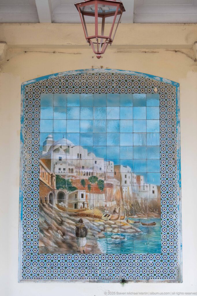 Ceramic wall mural of a waterfront city with boats and white buildings, framed by blue geometric tile patterns.