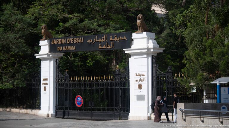 Monumental Jardin d’Essai du Hamma gate with bilingual black sign, white piers, sculptures, and pedestrians.