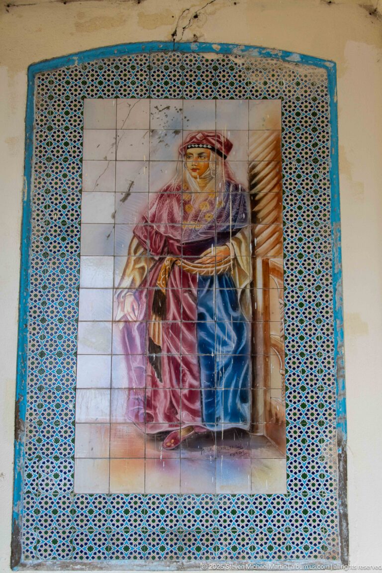 Weathered ceramic mural of a traditionally dressed standing figure bordered by turquoise and blue geometric tiles.