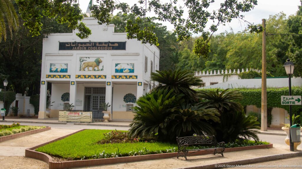 White Jardin Zoologique building with animal panels, cycads, lawn, bench, and shaded garden paths.