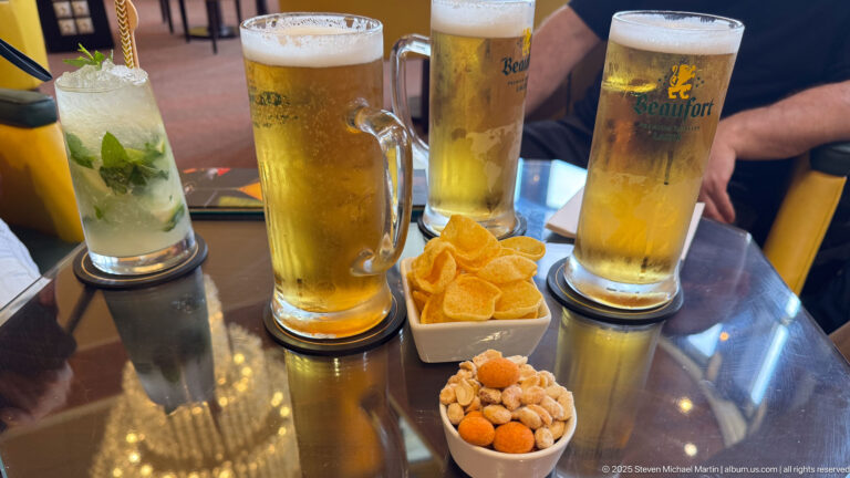 Three tall beers, a mint drink, potato chips, and peanuts arranged on a reflective café table.
