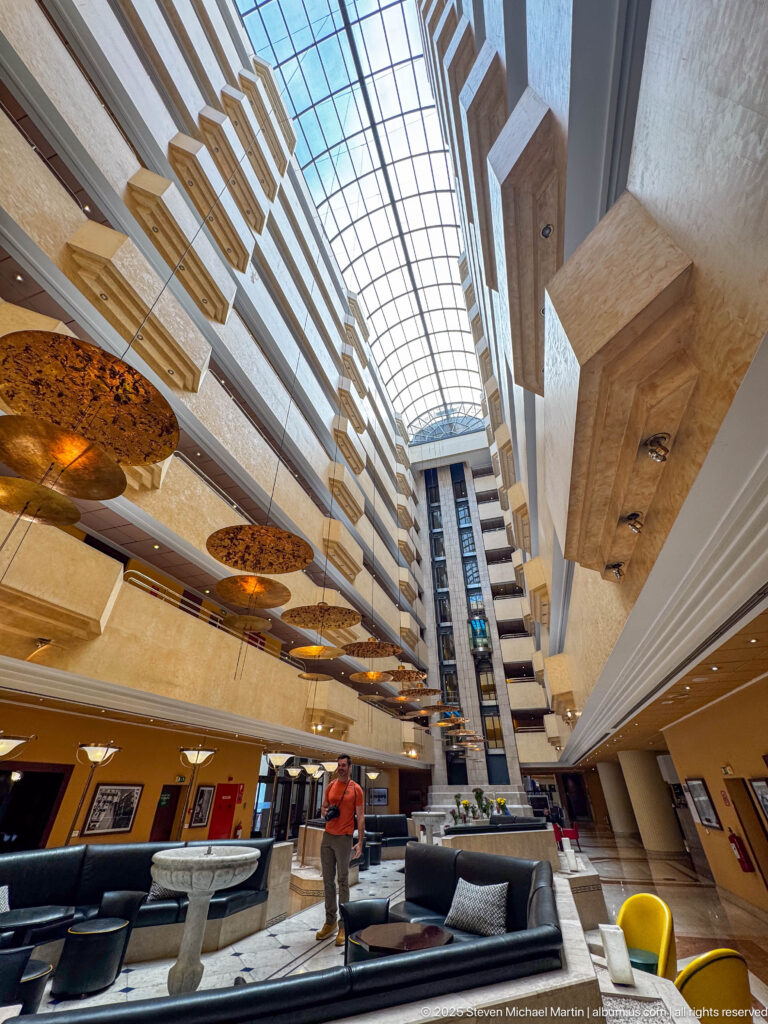 Tall modern atrium with stacked balconies, geometric projections, skylight, and lounge seating on the ground floor.