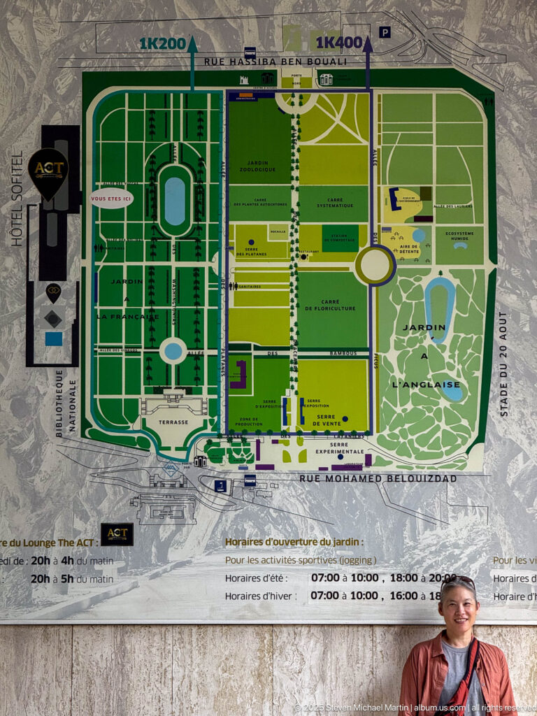 Large printed Jardin d’Essai du Hamma map with colored garden zones, paths, labels, and a seated visitor below.