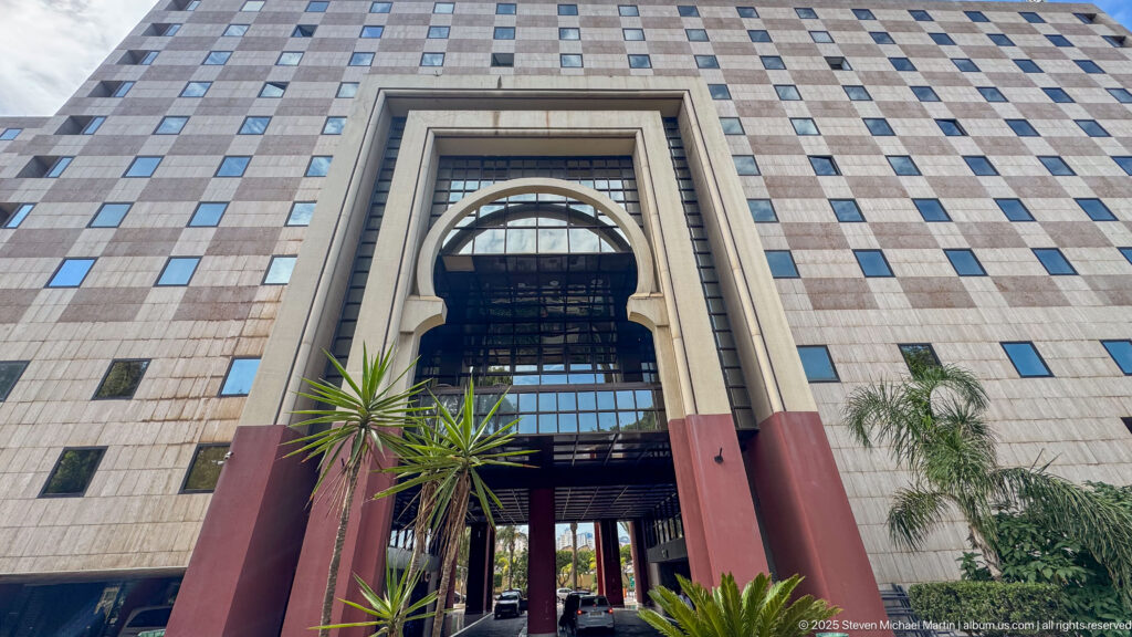 Tall stone-clad building with a large arched entrance, red-brown piers, gridded windows, and palms viewed from below.