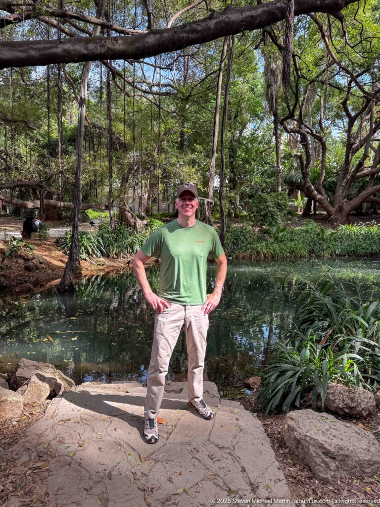 Man in a green shirt standing beside a shaded garden pond under spreading tree branches.