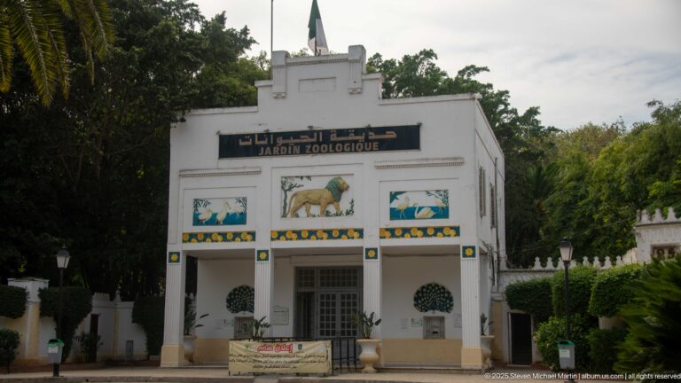White Jardin Zoologique entrance building with three animal panels above the doors and trimmed shrubs on both sides.