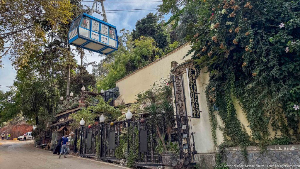 Cable-car cabin suspended above a landscaped street with garden fencing, vines, and pedestrians below.