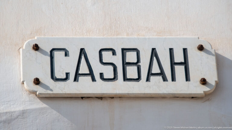 White rectangular CASBAH sign with black raised letters and rusted fasteners on a pale wall.