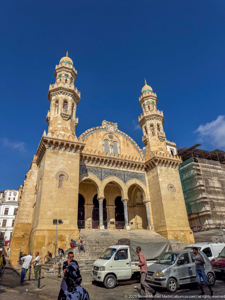 Large twin-towered pale-stone mosque above cars, scooters, pedestrians, and scaffolding along the street.