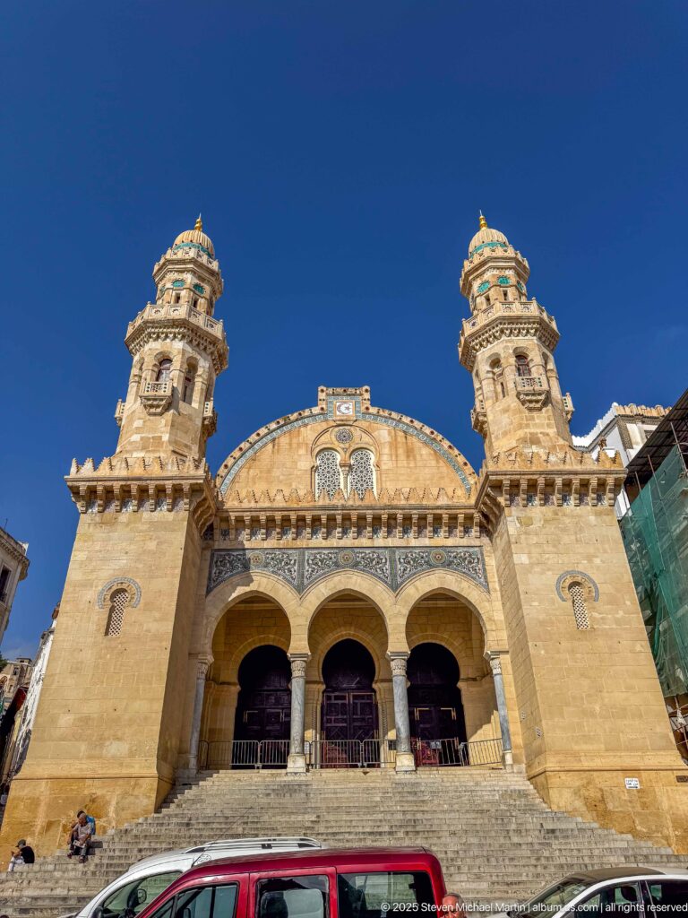 Frontal symmetrical view of pale-stone mosque with broad steps, three entrance arches, twin ornate towers, and turquoise accents.