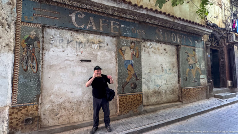 Visitor standing before a weathered Casbah café façade decorated with figurative mosaic panels and faded lettering.