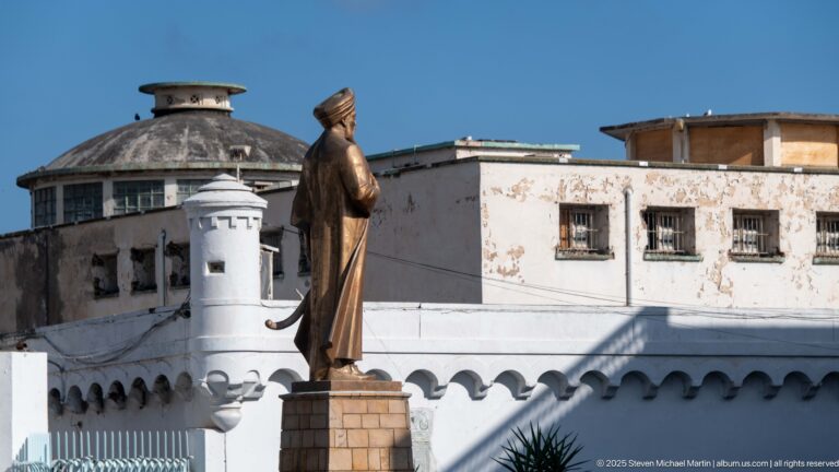 Bronze-colored statue of a robed man on a pedestal in front of white buildings and a small turret.