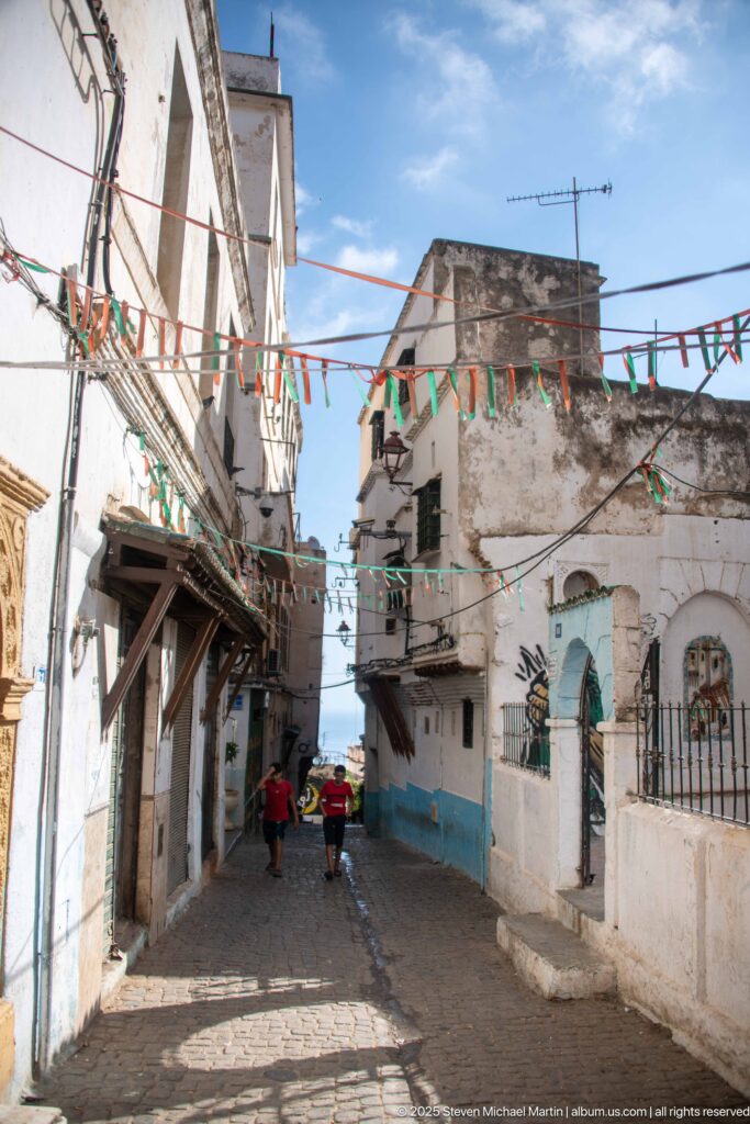 Narrow cobbled Casbah street with white buildings, pedestrians, balconies, overhead wires, and green-white-red bunting.