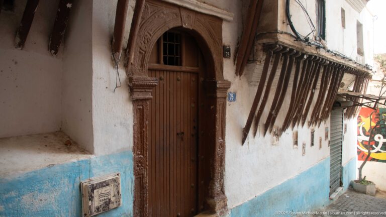 Carved arched wooden doorway in a white-and-blue Casbah façade beneath projecting dark timber braces.