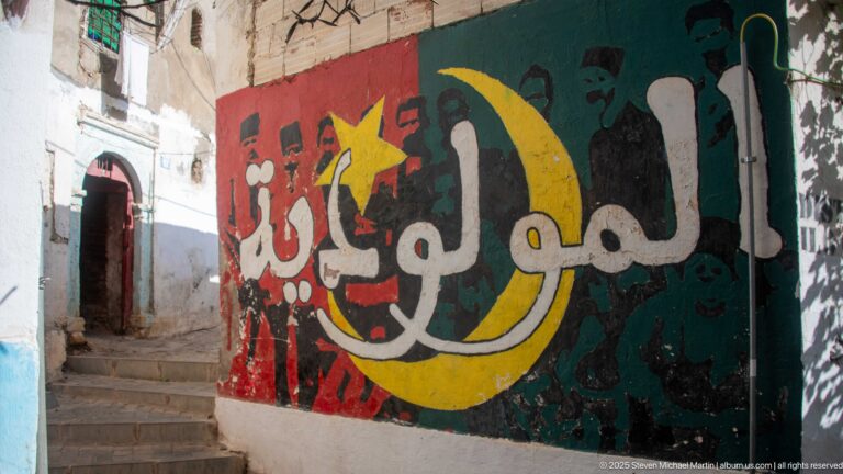Large Arabic calligraphy mural in red, green, white, and yellow beside steps in a narrow Casbah passage.