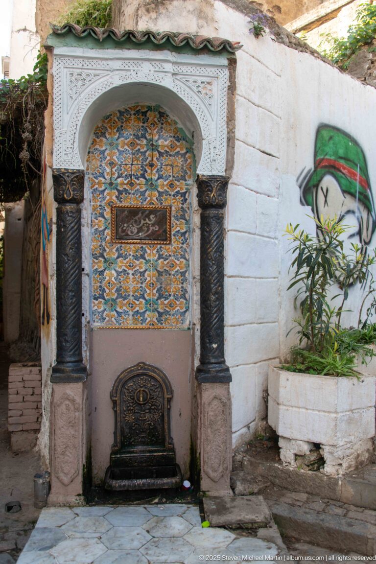 Decorative wall fountain with geometric tiles, white carved arch, dark columns, and a metal basin or spout.