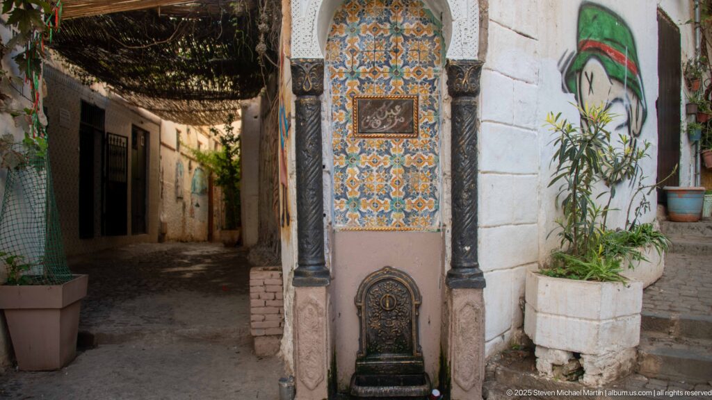Geometric-tiled wall fountain with dark columns and metal basin at the corner of a narrow Casbah passage.