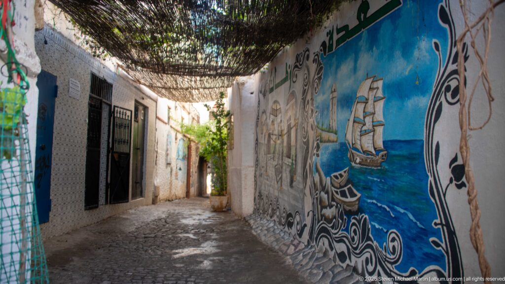 Blue mural of a multi-masted sailing ship on the wall of a narrow shaded Casbah alley.