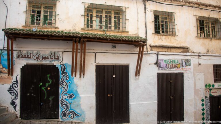 Weathered white Casbah façade with three dark doors, barred windows, green-tiled canopy, Arabic signage, and painted ornament.