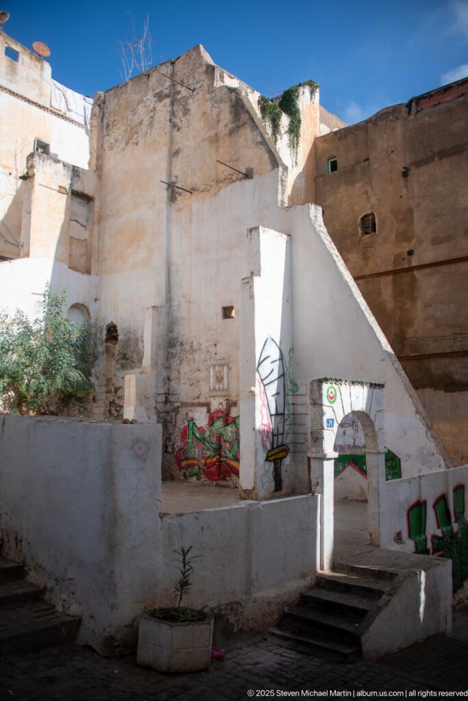 Whitewashed stepped courtyard with wall murals, planters, green seating, and tall weathered masonry above.