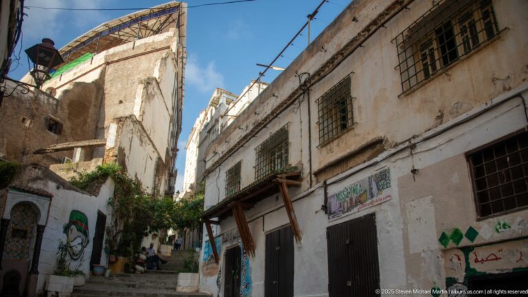 Narrow Casbah lane between tall weathered buildings with barred windows, patched plaster, signs, and painted wall decoration.