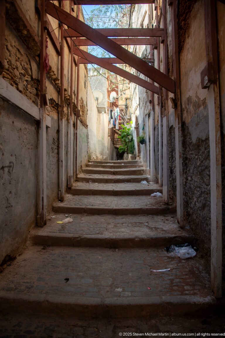 Steep narrow Casbah stairway with weathered walls, overhead cross-bracing, pipes, and daylight at the upper end.