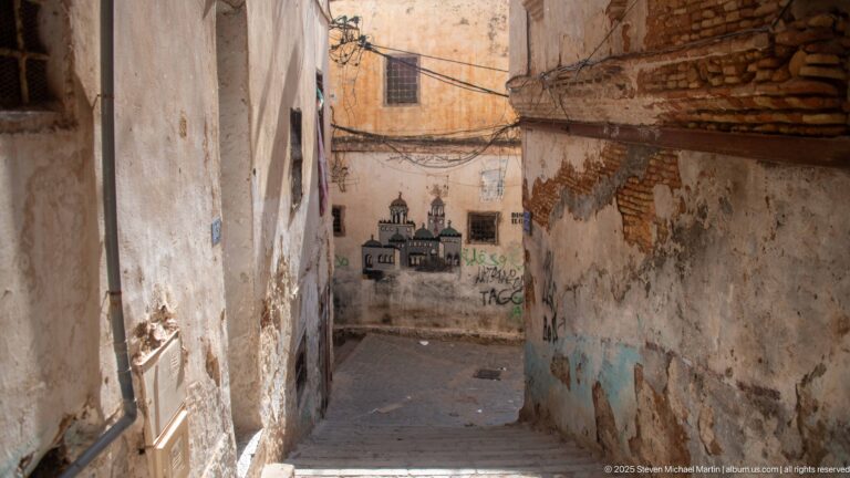 Descending narrow Casbah alley with peeling plaster, exposed brick, graffiti, overhead wires, and an architectural mural ahead.