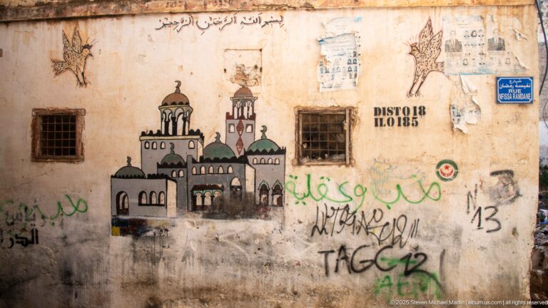 Weathered wall painted with a domed mosque-like building, surrounded by Arabic writing, graffiti, notices, and barred windows.