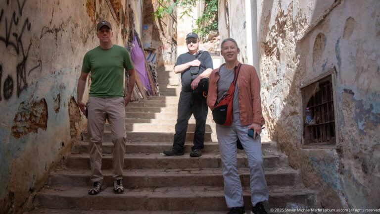 Three visitors standing on steep stone steps in a narrow Casbah alley with weathered walls and plants.