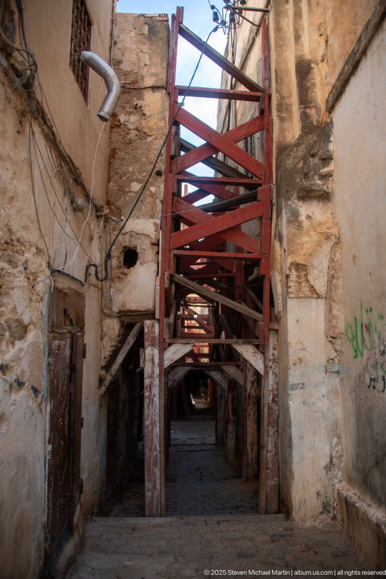 Narrow Casbah passage filled with heavy red cross-bracing between weathered stone and plaster walls.