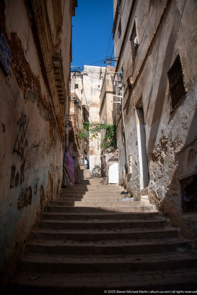 Broad stone stairway rising between weathered Casbah buildings, with overhead wires and bright sunlight above.